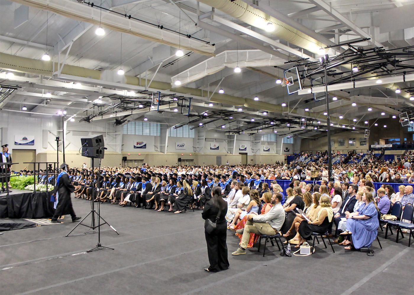 A large crowd gathers in a gymnasium for a graduation ceremony. As Buffalo Rock Company expands to Loxley, excitement fills the air, with graduates in caps and gowns sitting in rows and families filling the audience seats near the stage and microphone.